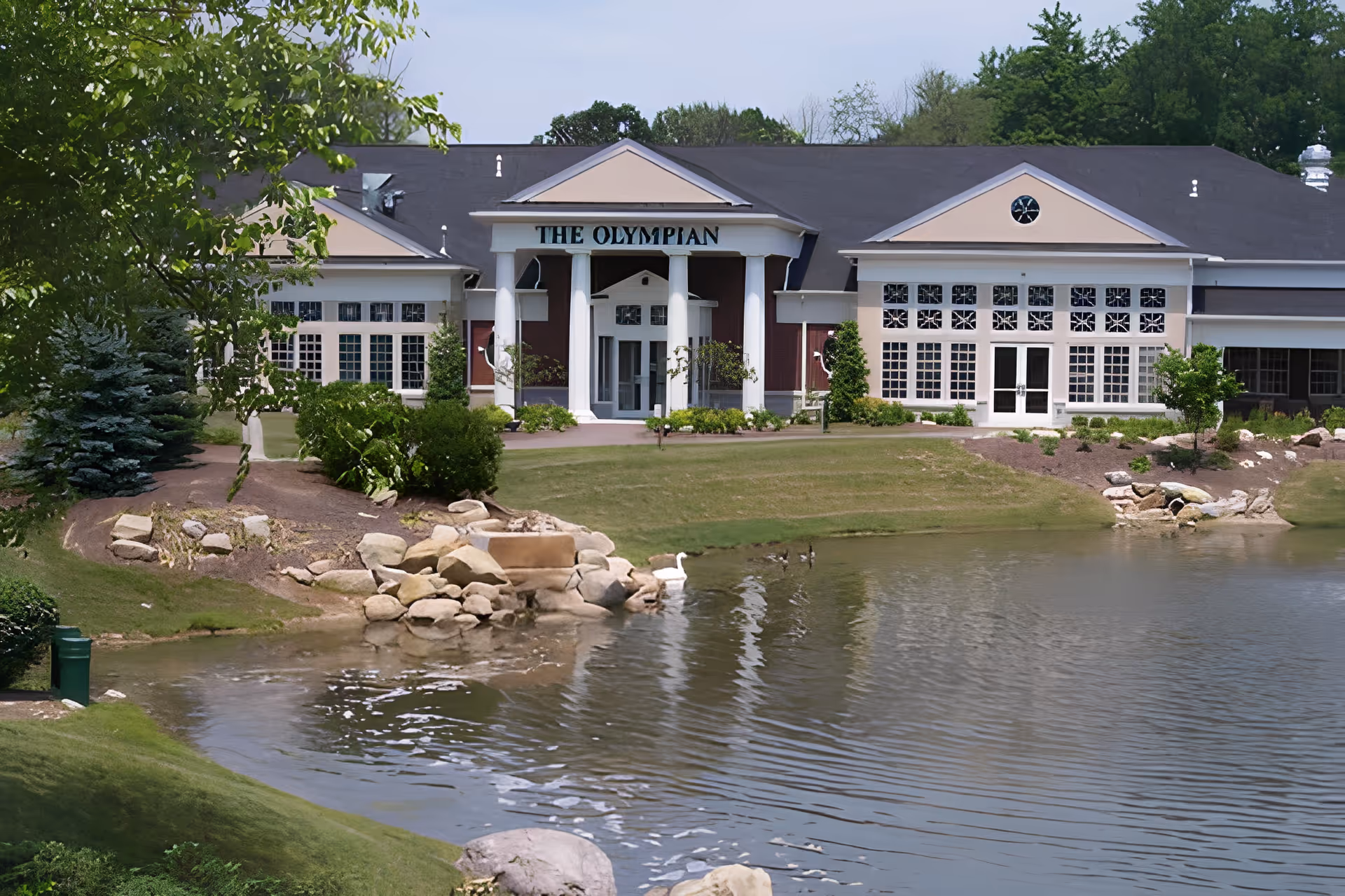 Exterior view of The Olympian building at Gaitanaros Commons, featuring a large pond in the foreground, landscaped greenery, and a building with white columns and multiple windows.