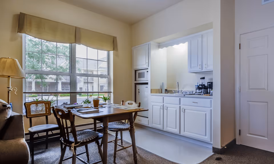 A bright room with a wooden dining table and four chairs, a large window with blinds and a beige valance, a floor lamp, and a small kitchen area with white cabinets, a microwave, a sink, and various kitchen appliances.