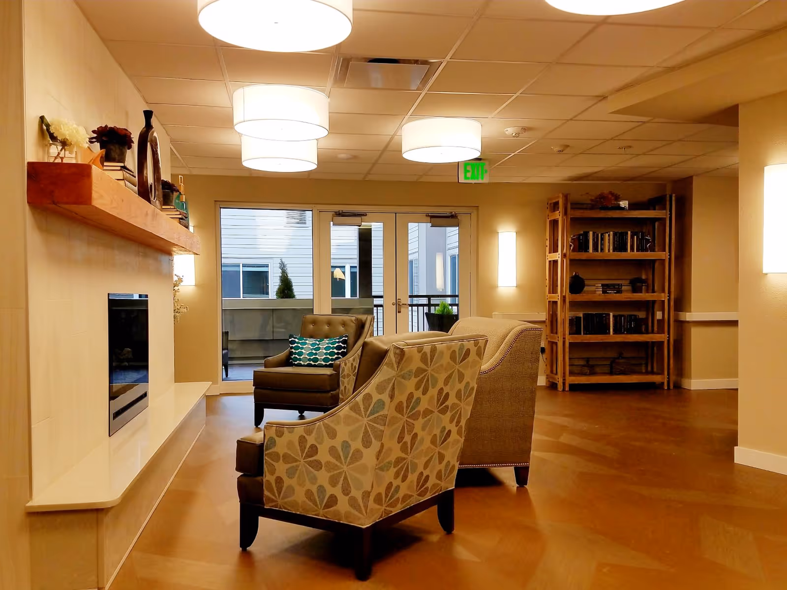 Cozy senior living lounge with patterned armchairs, a wall fireplace, bookshelf, and ceiling lights near glass doors to an outdoor area.