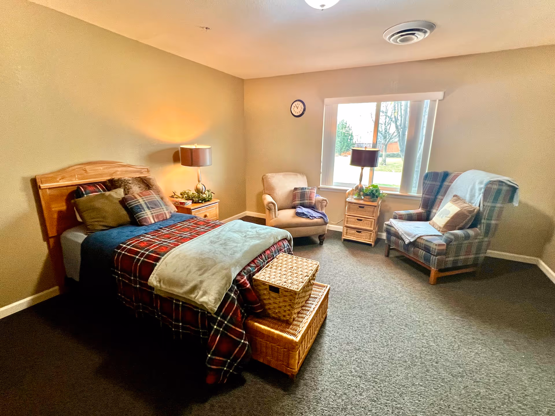 A cozy bedroom in Aspen House featuring a single bed with plaid bedding and multiple pillows. Next to the bed is a wooden nightstand with a lamp and decorative greenery. Across from the bed are two armchairs, one beige and one plaid, with a small wicker drawer unit and a lamp between them. A large window lets in natural light, and the room has beige walls and carpeted flooring.