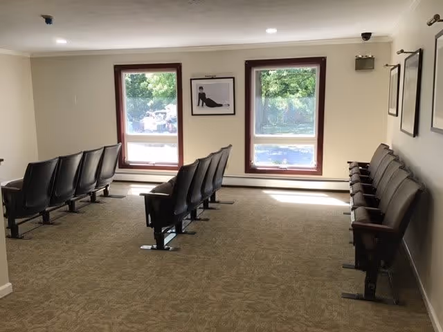 A small room with three rows of black theater-style seats facing two windows with brown frames. The room has beige walls and carpeted floor. There are framed pictures on the walls and natural light coming through the windows.