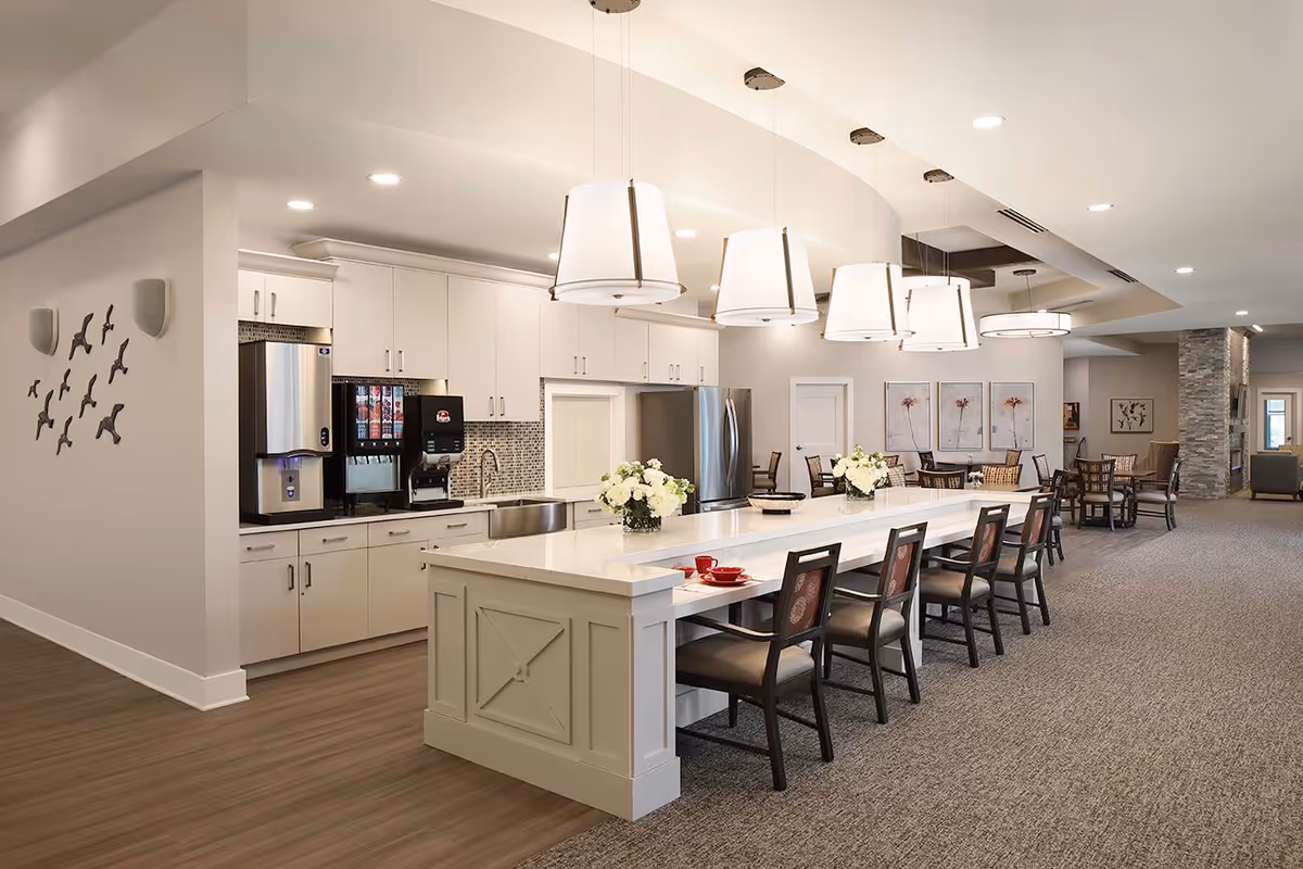 A modern senior living facility kitchen and dining area with a long white island counter featuring six chairs. The kitchen has white cabinets, stainless steel appliances, and a beverage dispenser. The dining area includes multiple tables and chairs with floral artwork on the walls. The space is well-lit with pendant lights above the island and recessed ceiling lights.
