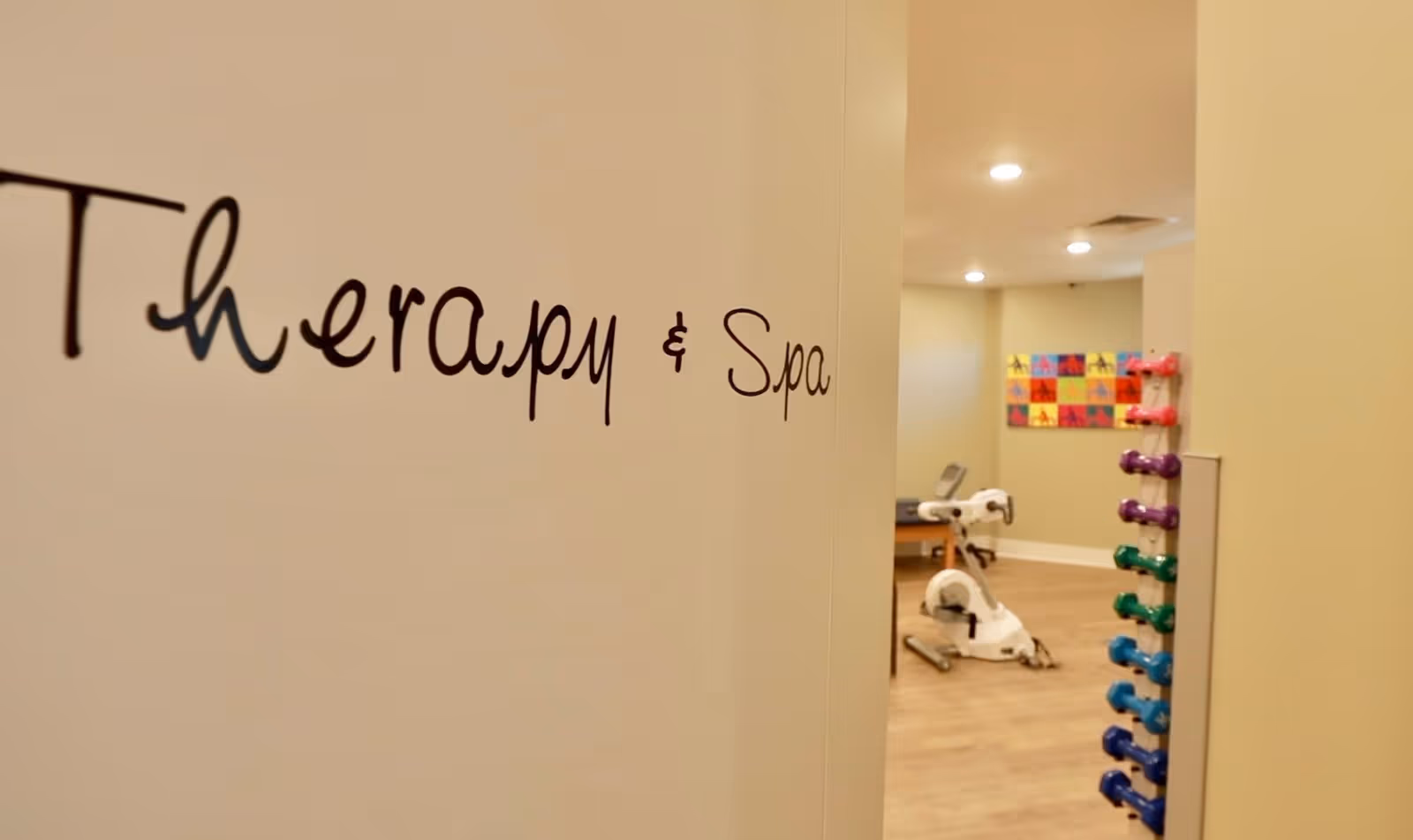 Interior view of a therapy and spa room in a senior living facility, showing a stationary exercise bike, a rack of colorful dumbbells, and a colorful wall art piece in the background.