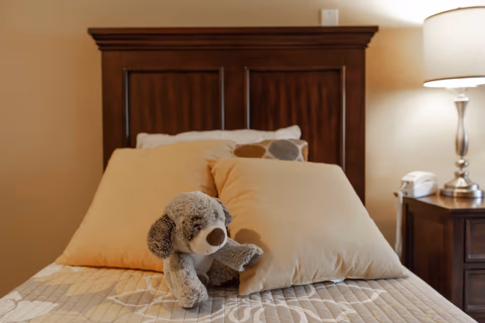 A neatly made bed with a wooden headboard, beige pillows, and a gray stuffed dog toy placed on top. To the right of the bed is a wooden nightstand with a silver lamp and a white telephone.