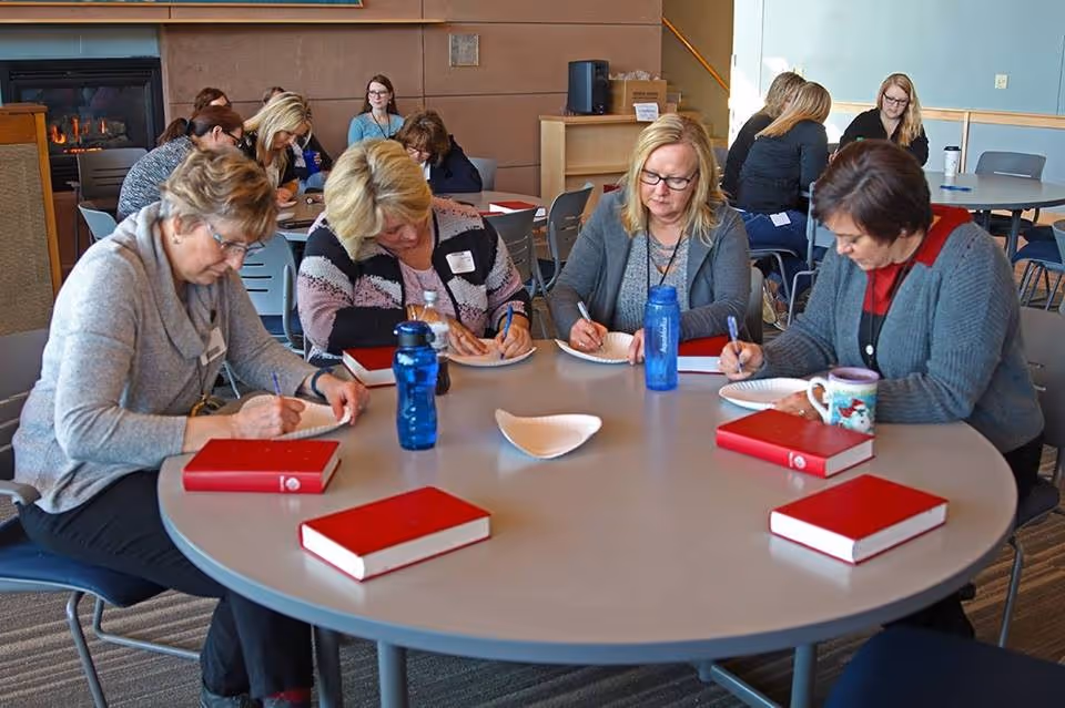 A group of women sitting around a large round table in a common area, writing on paper plates. There are several red books on the table, along with water bottles and a coffee mug. In the background, more people are seated at tables, and a fireplace is visible on the left side of the room.