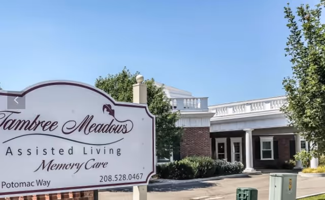 Outdoor view of Tambree Meadows Assisted Living facility showing the entrance area with a white sign in the foreground that reads 'Tambree Meadows Assisted Living Memory Care' along with a phone number and street name. The building has brick walls, white columns, and a clear blue sky above.