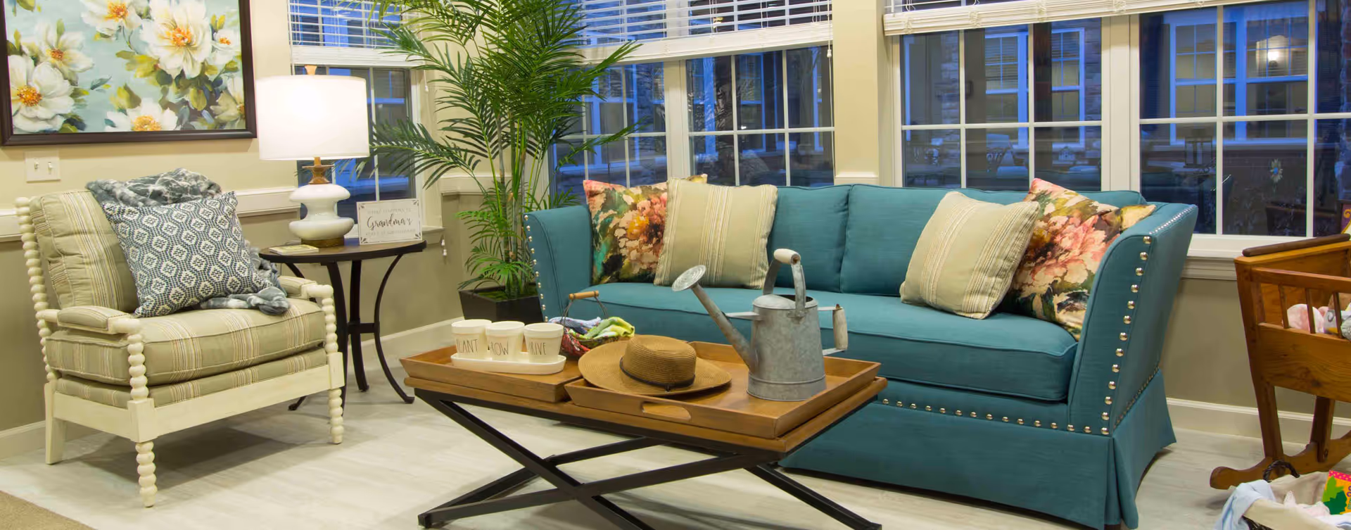 A cozy living room area with a teal sofa adorned with floral and striped pillows, a wooden coffee table with a watering can, a hat, and three white pots labeled Plant, Grow, Thrive. To the left is a cushioned armchair with patterned pillows and a throw blanket, next to a side table with a white lamp and a small sign. Large windows with blinds are in the background, and a tall green plant is placed near the corner.