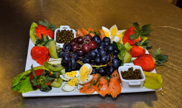 A rectangular white plate with an assortment of food including strawberries, sliced kiwi, hard-boiled egg quarters, smoked salmon, green lettuce leaves, capers in small white square dishes, and a cluster of red and black grapes in the center, all arranged on a metallic surface.