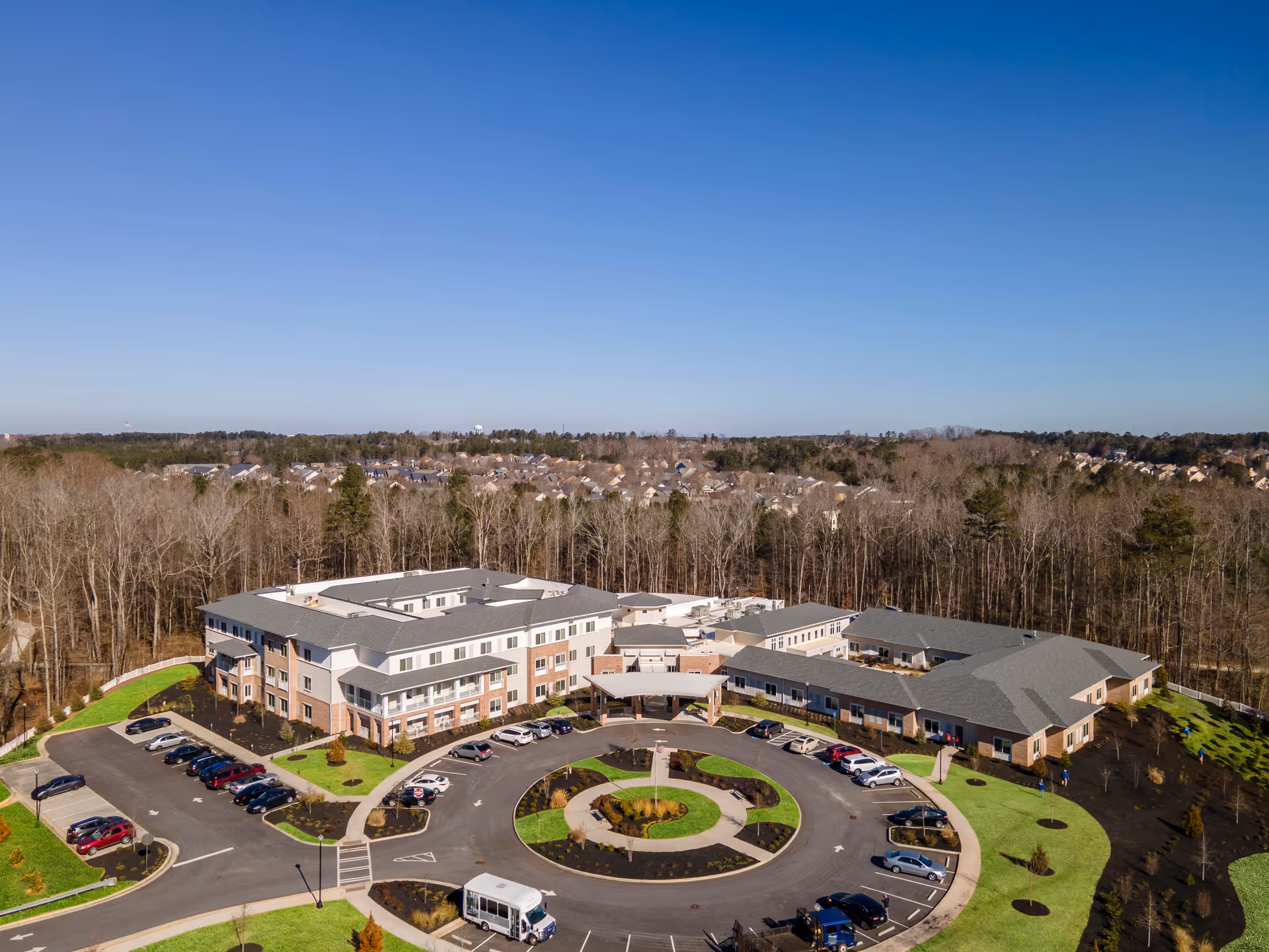 Aerial view of Westhill Newnan Crossing senior living facility showing a large, multi-wing building with a circular driveway and parking lot surrounded by green lawns and trees, with a residential neighborhood and forest in the background under a clear blue sky.