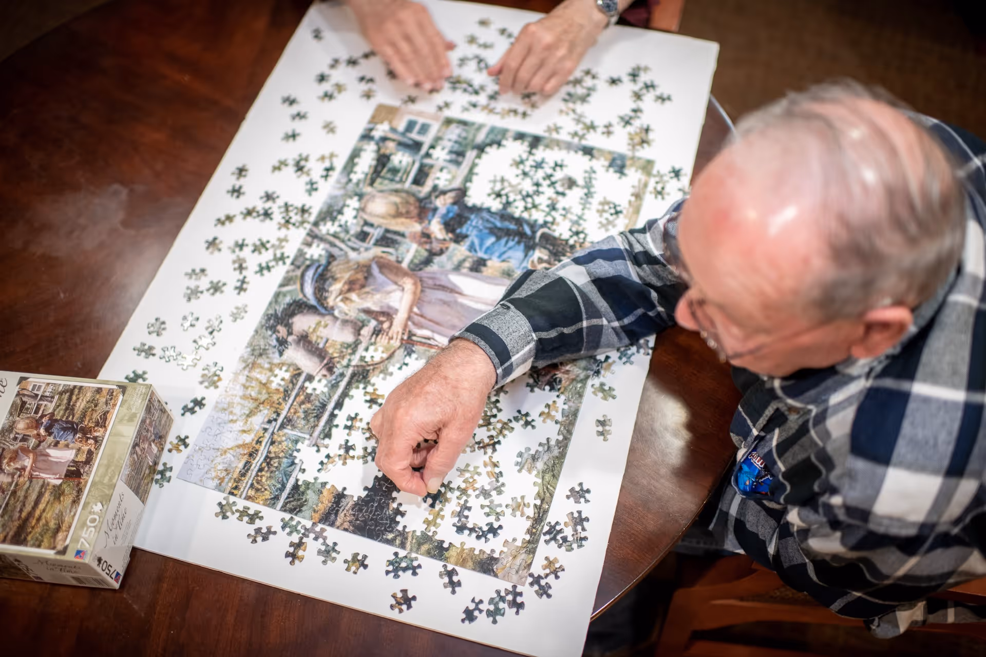 An elderly man wearing glasses and a plaid shirt is working on assembling a jigsaw puzzle on a wooden table. The puzzle depicts a vintage scene with people and a horse. Another person's hands are visible at the top of the image, also engaged with the puzzle pieces.