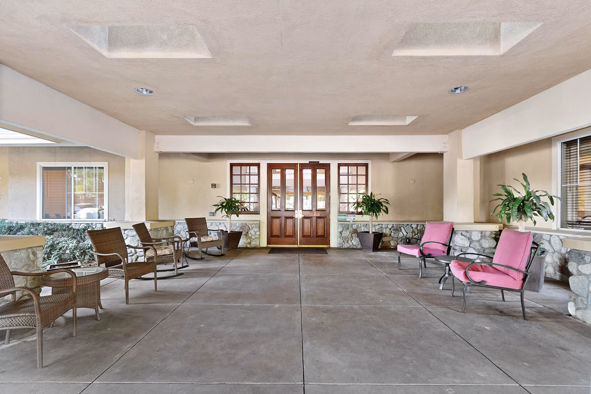 Covered outdoor seating area at Claremont Place with wicker chairs and pink cushioned metal chairs arranged along the sides, potted plants near the stone wall, and double wooden doors at the far end.