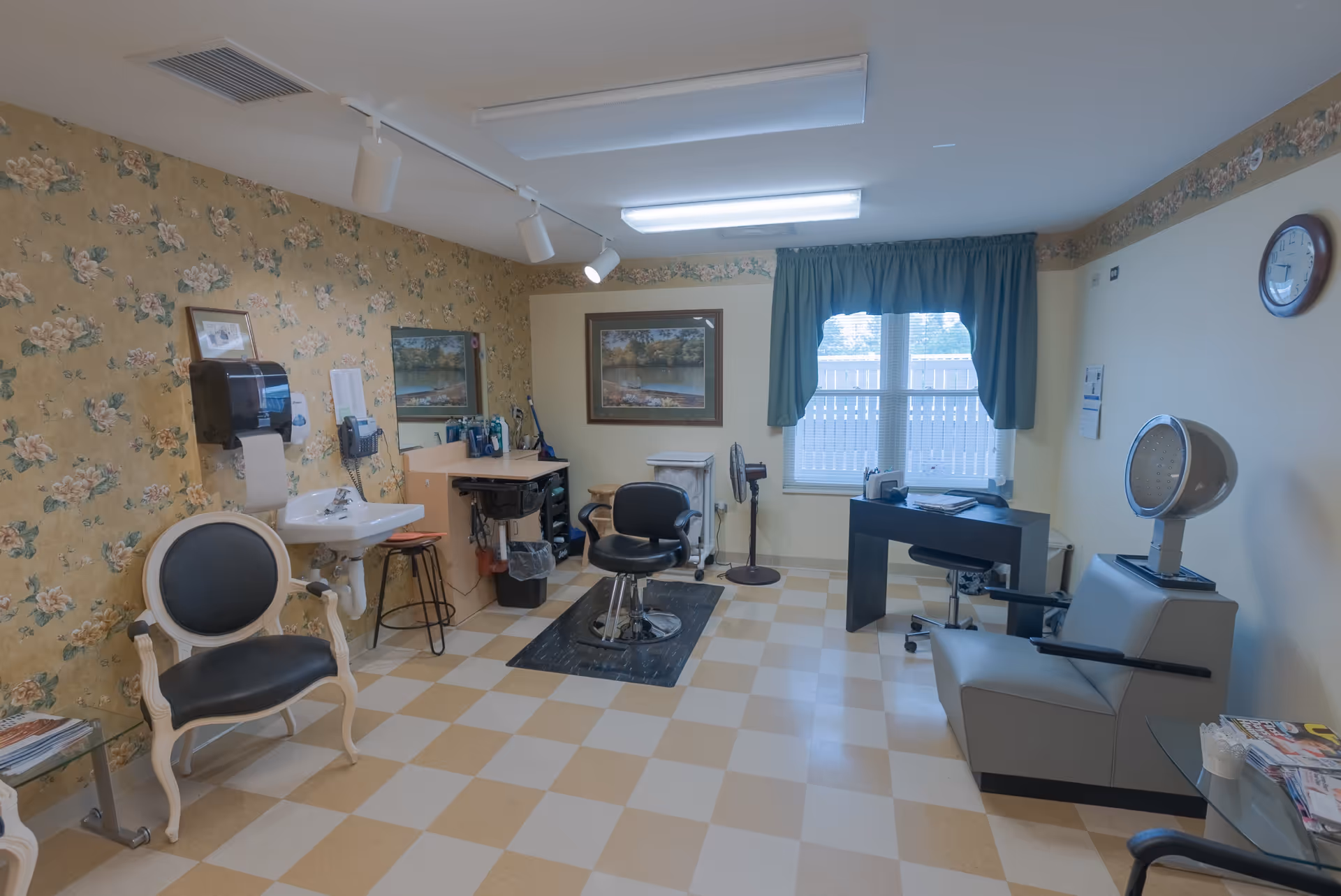 Interior view of a hair salon room in a senior living facility with a floral wallpapered wall, a black salon chair in front of a mirror and sink, a vintage hair dryer chair, a desk with a chair near a window with green curtains, and a clock on the wall.