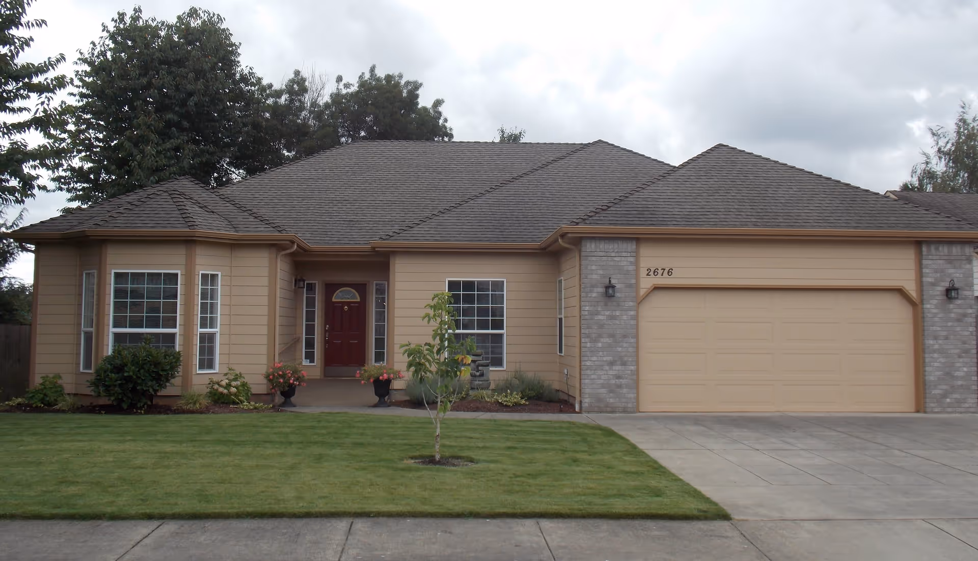 Front exterior view of a single-story house with beige siding, a dark brown front door, two large windows, a two-car garage, a small tree in the front yard, and a well-maintained lawn under a cloudy sky.