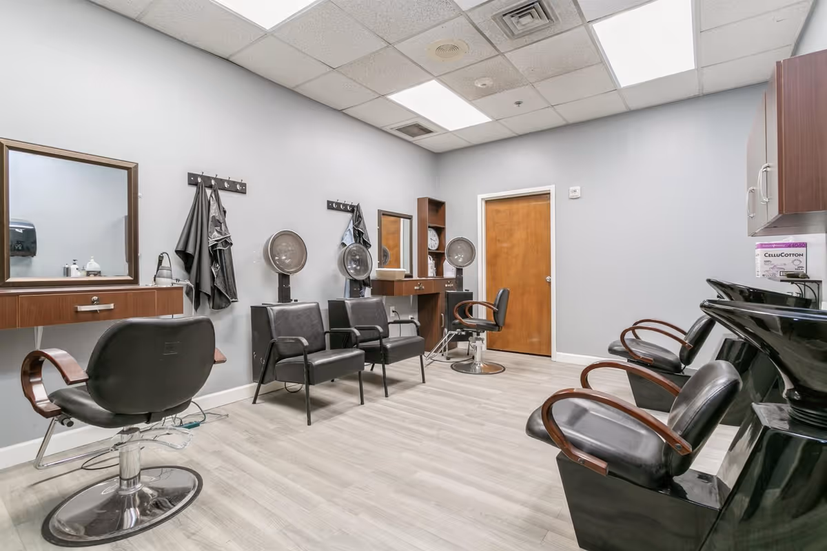 Interior view of a hair salon room with several black salon chairs, hair drying stations, mirrors on the wall, and a wooden door. The room has light gray walls, a light wood floor, and ceiling lights.