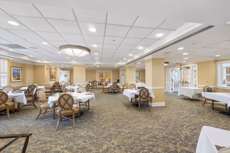 A spacious dining room with multiple tables covered in white tablecloths and surrounded by cushioned chairs. The room has beige walls, carpeted floors, recessed lighting, and large windows allowing natural light. There are framed pictures on the walls and a decorative ceiling light fixture.