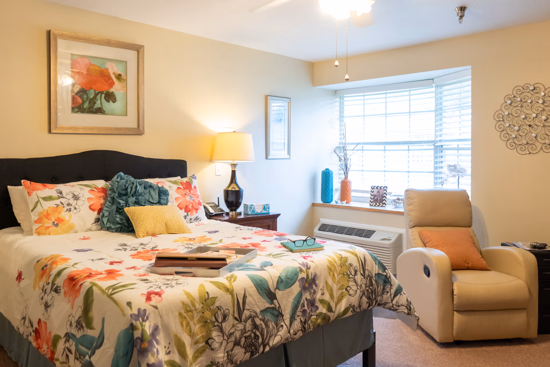 A cozy bedroom with a bed covered in floral bedding and multiple pillows. There is a bedside table with a lamp, phone, and tissue box. A comfortable beige recliner chair with an orange pillow sits near a window with white blinds. Decorative items and framed artwork adorn the walls and window sill.