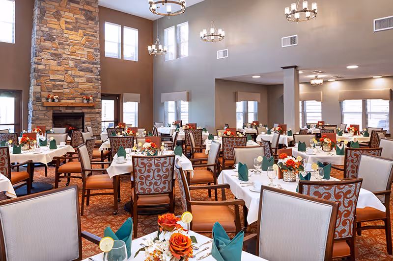 A spacious dining room with multiple tables covered in white tablecloths, each set with green folded napkins, glassware, and floral centerpieces featuring orange and red flowers. The room has high ceilings with large windows allowing natural light, stone fireplace on one side, and elegant chandeliers hanging from the ceiling.