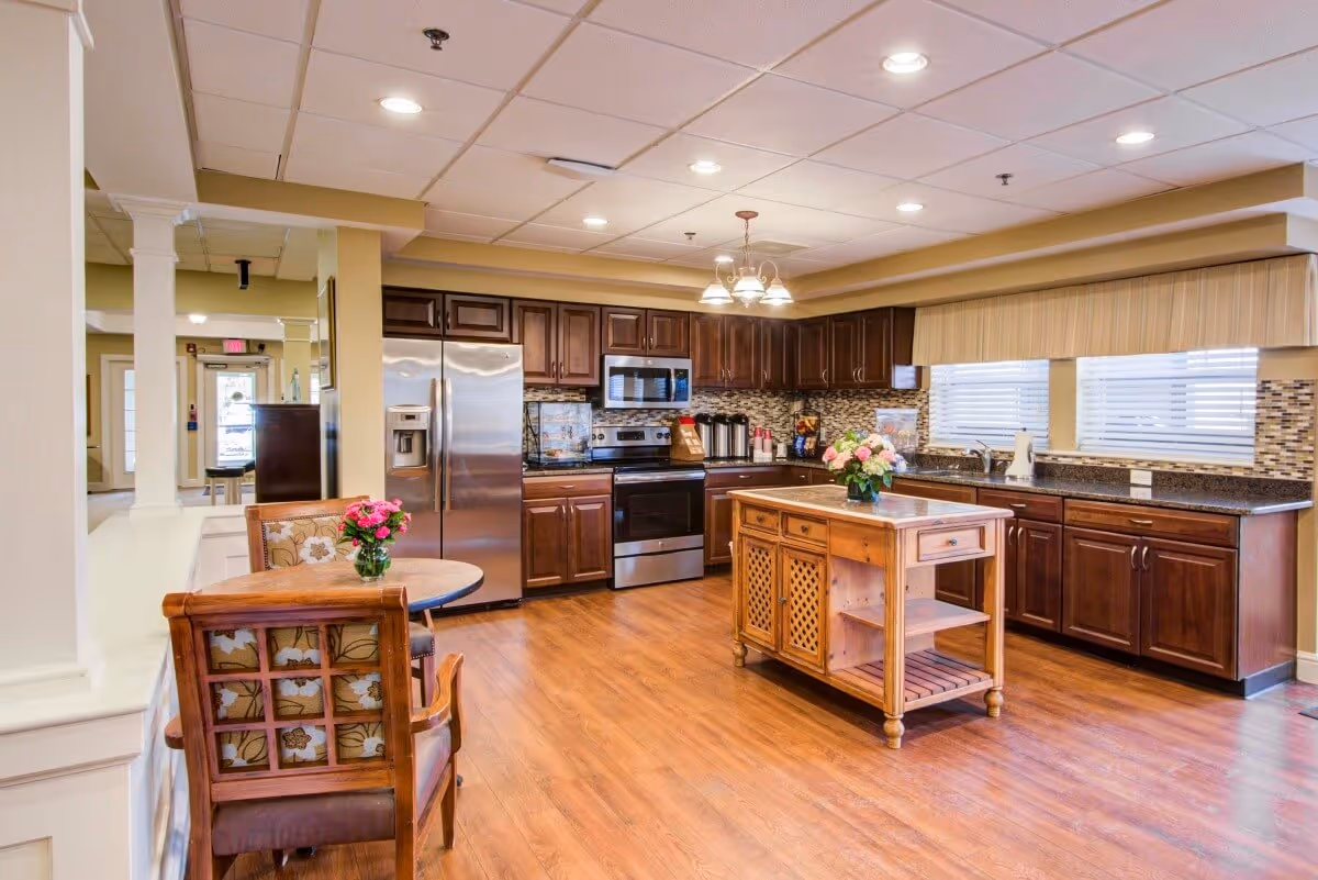 A spacious kitchen area with wooden flooring and dark wood cabinets. The kitchen features stainless steel appliances including a refrigerator, oven, and microwave. There is a wooden island in the center with a flower vase on top. To the left, there is a small round table with two cushioned wooden chairs, also decorated with a flower vase. The room is well-lit with recessed ceiling lights and a hanging light fixture above the island. Large windows with blinds allow natural light into the space.