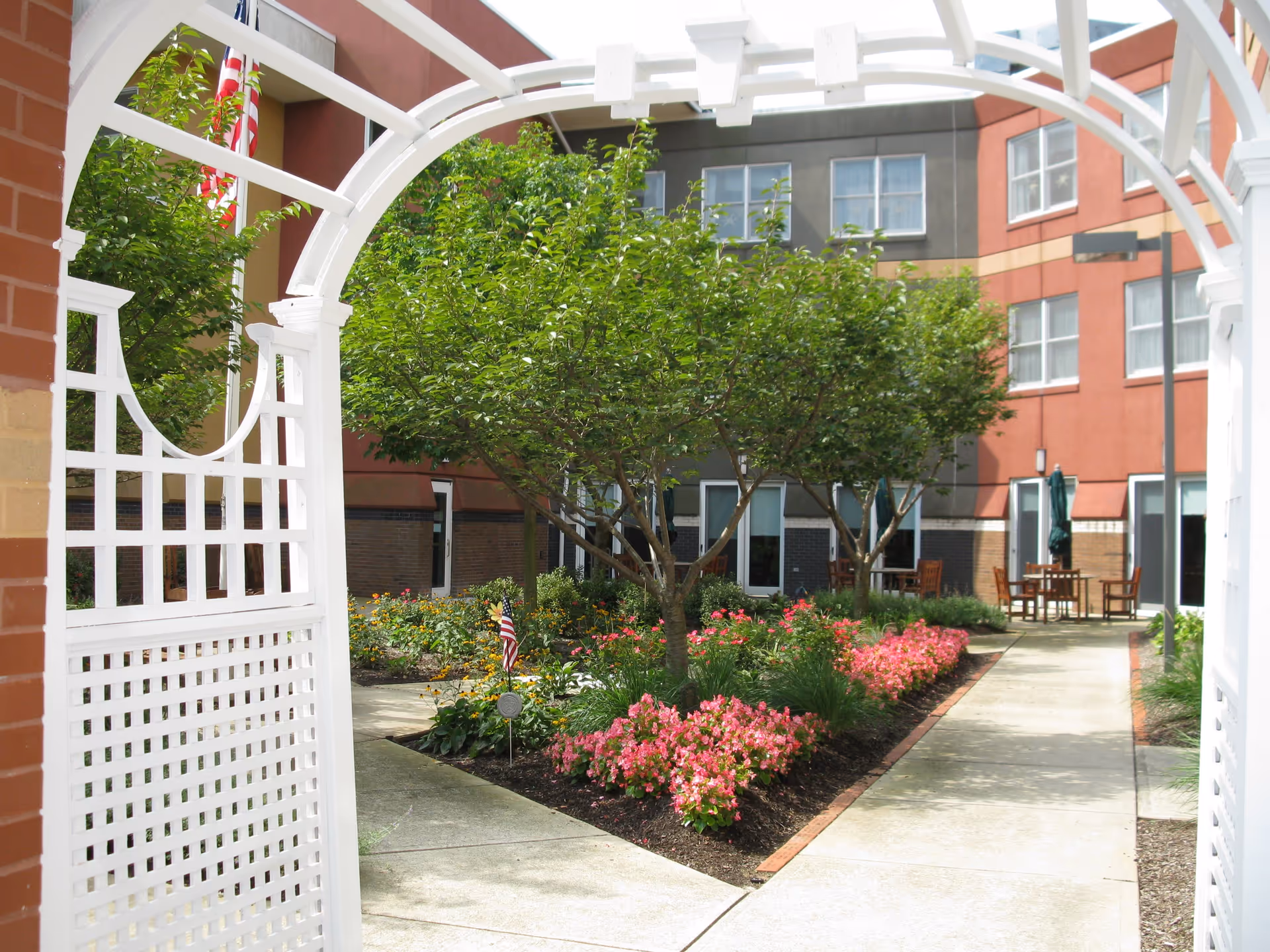 View through a white wooden archway into a courtyard garden with green trees, pink flowers, and paved walkways. Surrounding the garden are multi-story buildings with windows and outdoor seating areas with tables and chairs.