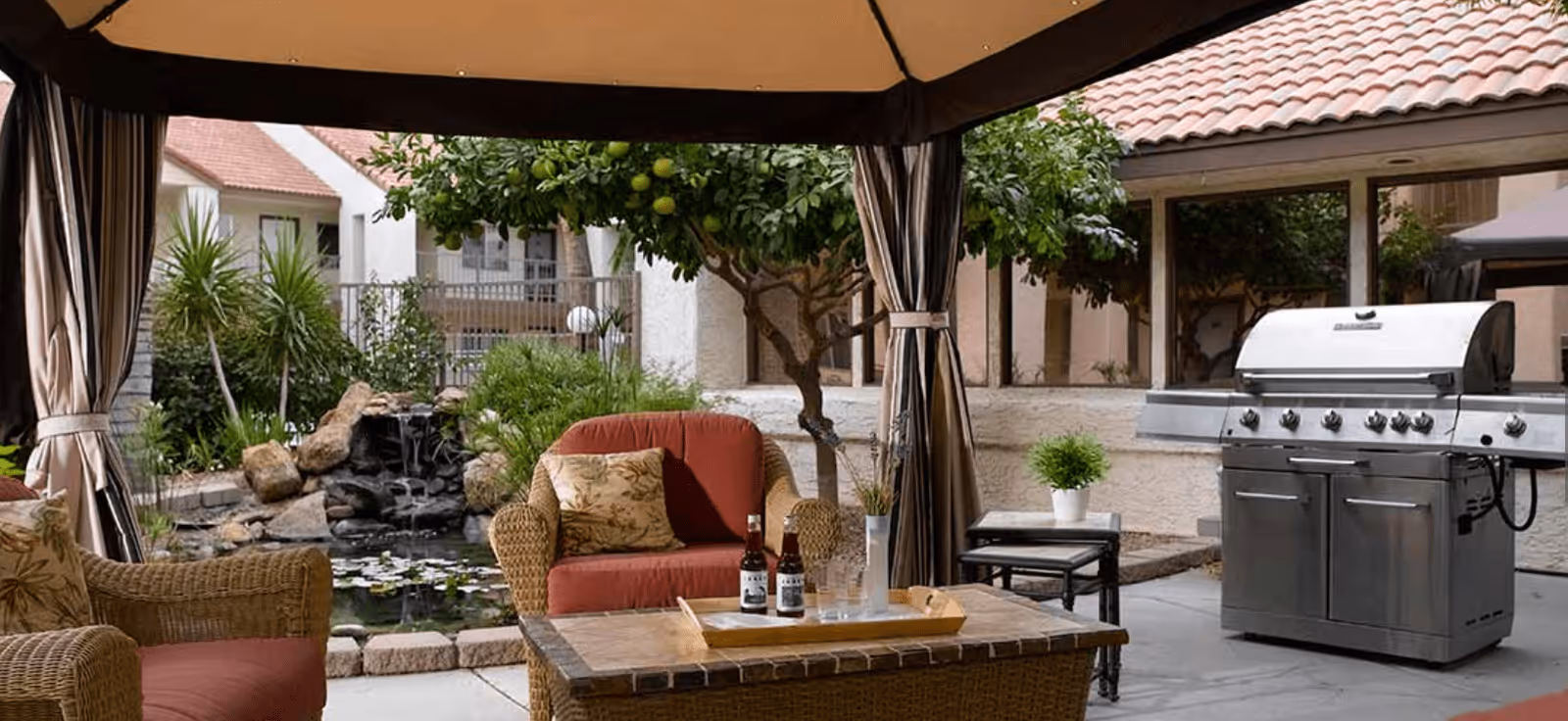 Outdoor patio area with wicker furniture including a cushioned armchair and loveseat, a coffee table with drinks, a large stainless steel grill, and a small side table with a potted plant. The space is shaded by a canopy and surrounded by greenery, including a small tree and a rock waterfall feature with a pond.