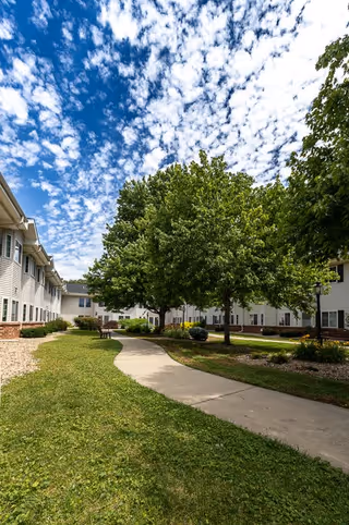 A paved walkway curves through a grassy courtyard with green trees and shrubs, surrounded by two-story beige buildings under a partly cloudy blue sky.