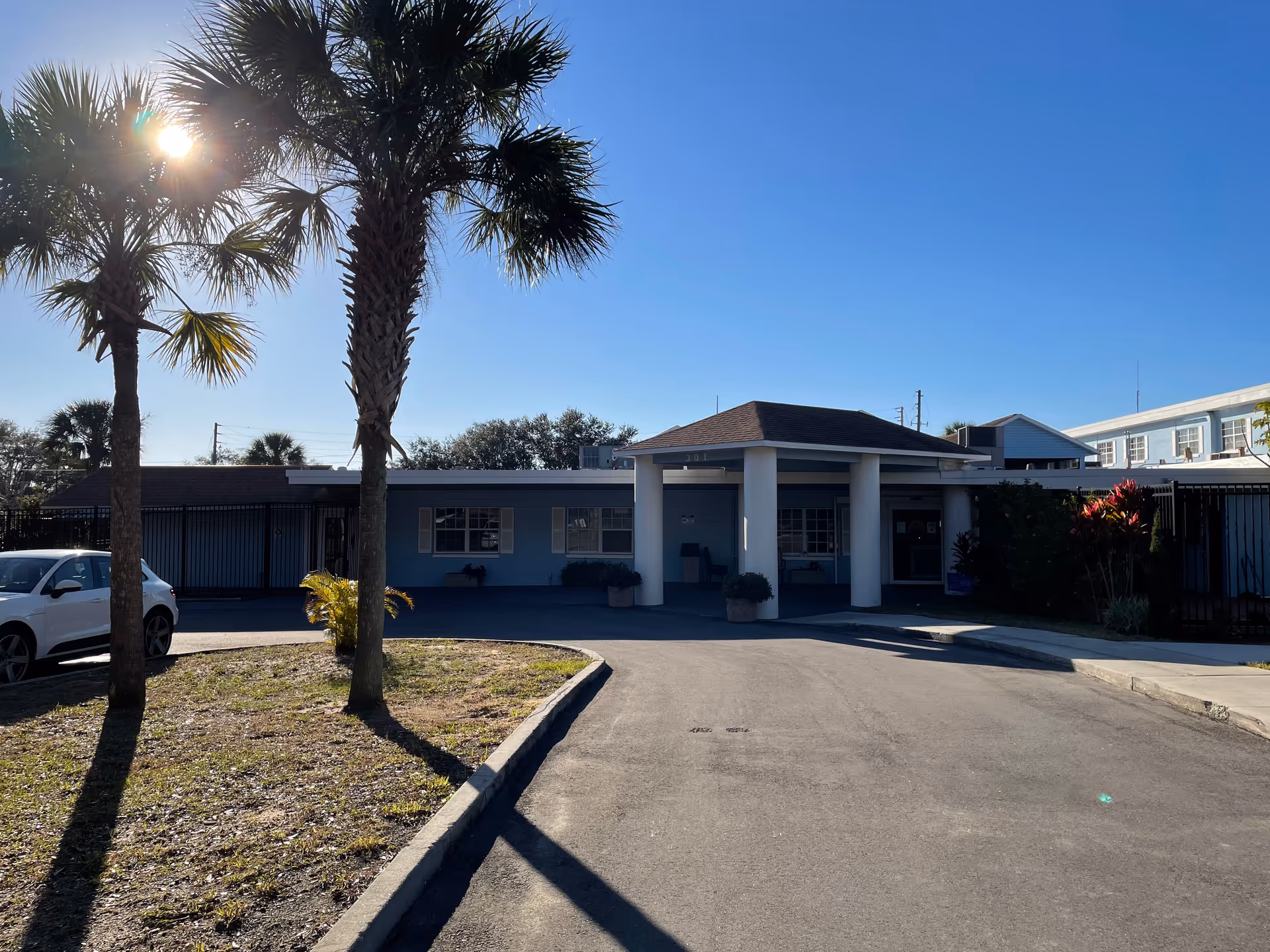 Front exterior of a single-story assisted living building with a covered entrance, palm trees, and a driveway under a clear blue sky.