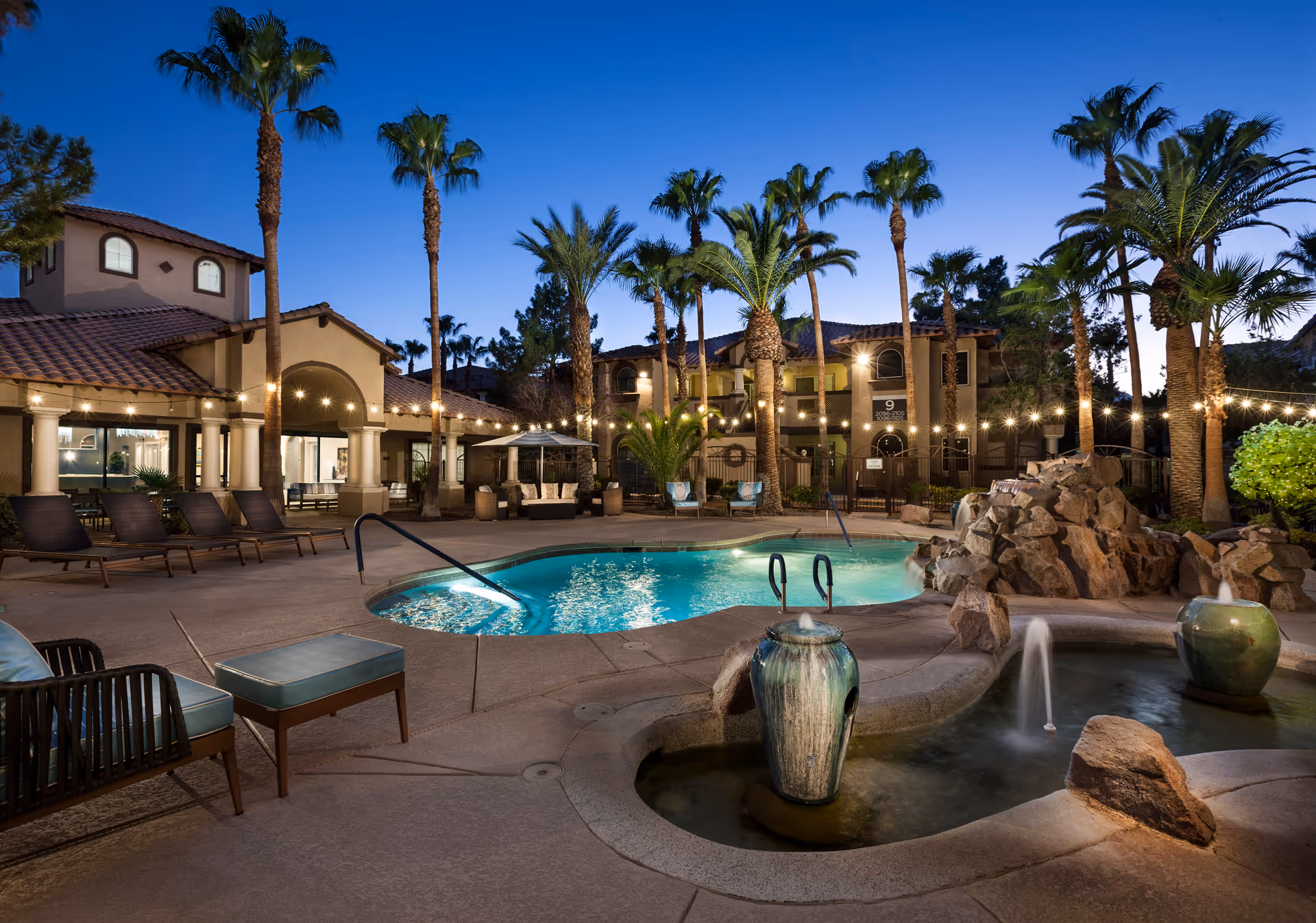 Outdoor pool area at dusk with palm trees, lounge chairs, a small fountain, and string lights illuminating the space. The building with arched windows and tiled roof surrounds the pool area.