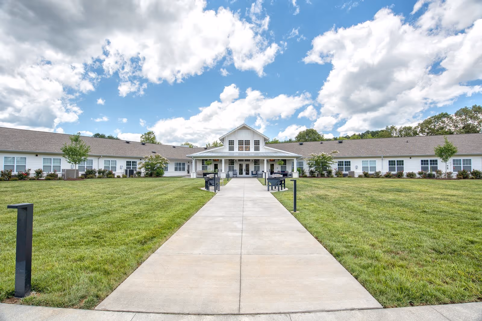 Wide front view of a single-story senior living facility building with a large green lawn and a concrete walkway leading to the entrance under a partly cloudy sky.