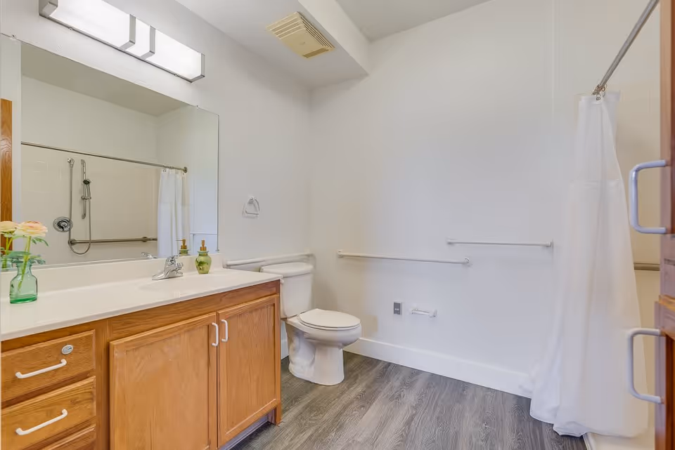 A clean and spacious bathroom with a wooden vanity featuring a white countertop and a large mirror above it. There is a toilet next to the vanity and a shower area with a white curtain and grab bars for safety. The floor has a wood-like finish and the walls are painted white.