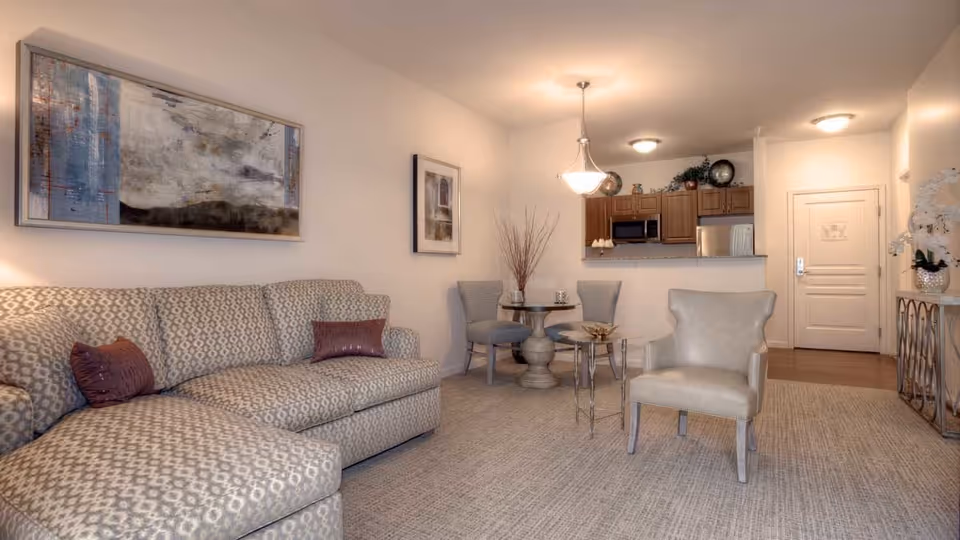A cozy living room area with a patterned sectional sofa adorned with two purple pillows, a single beige armchair, and a small glass side table. In the background, there is a round dining table with two chairs, a kitchen with wooden cabinets, a microwave, and a refrigerator. The walls are decorated with framed artwork, and the room is softly lit by ceiling lights and a hanging light fixture.