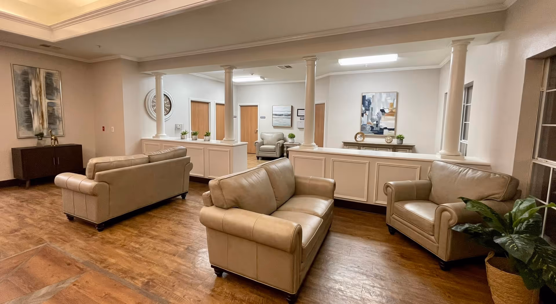 A bright communal lounge with beige leather sofas and armchairs, decorative columns, artwork, and wood flooring.