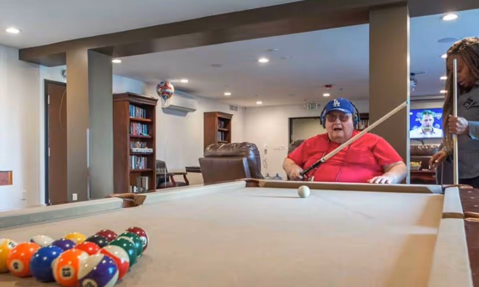 An elderly man wearing a blue cap and red shirt is playing pool indoors, holding a cue stick and aiming at the cue ball on a pool table. A woman stands nearby holding another cue stick. The room has bookshelves, chairs, and a television in the background.