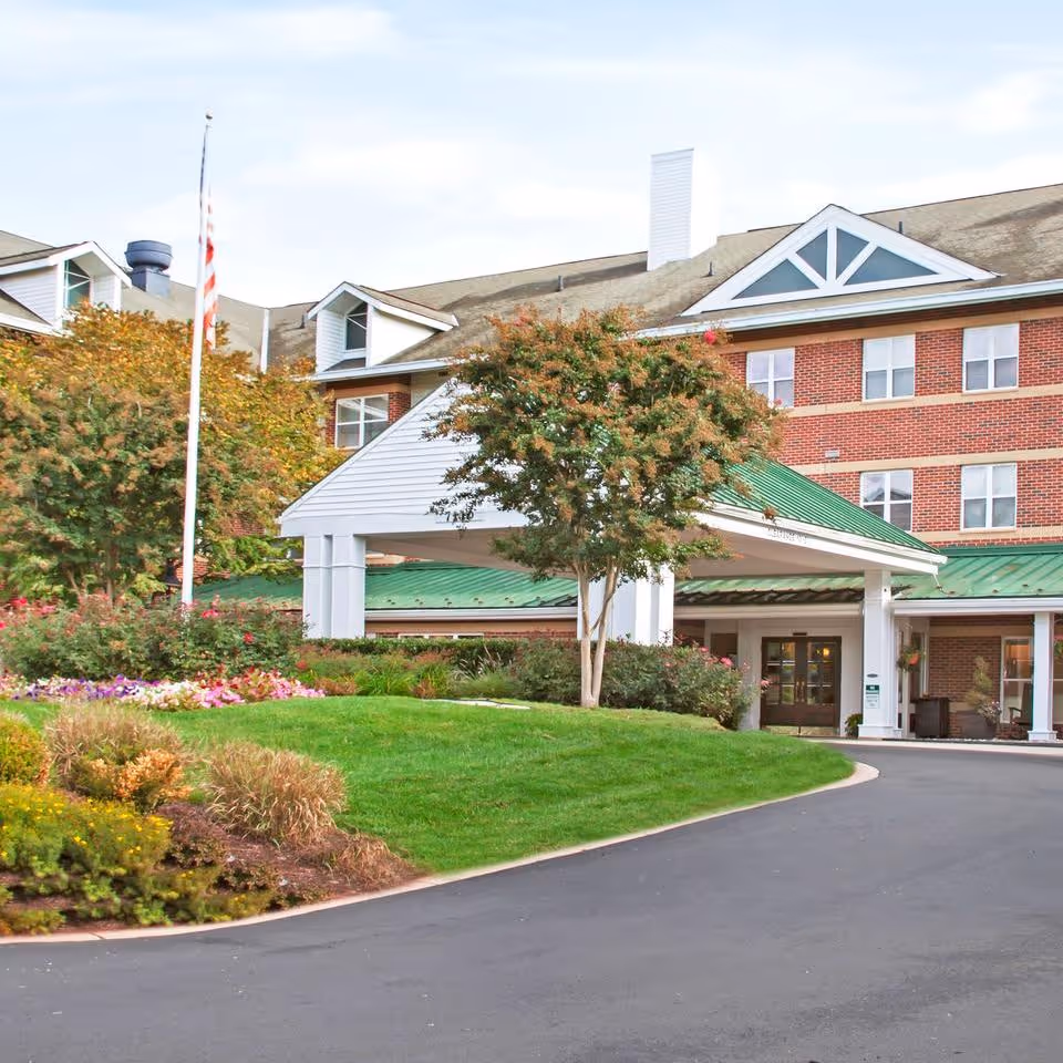 Exterior view of Brighton Gardens of Columbia, showing a multi-story brick building with white trim and a green metal roof over the entrance. There is a curved driveway leading to the entrance, landscaped with green grass, bushes, flowers, and trees. An American flag is visible on a flagpole near the entrance.