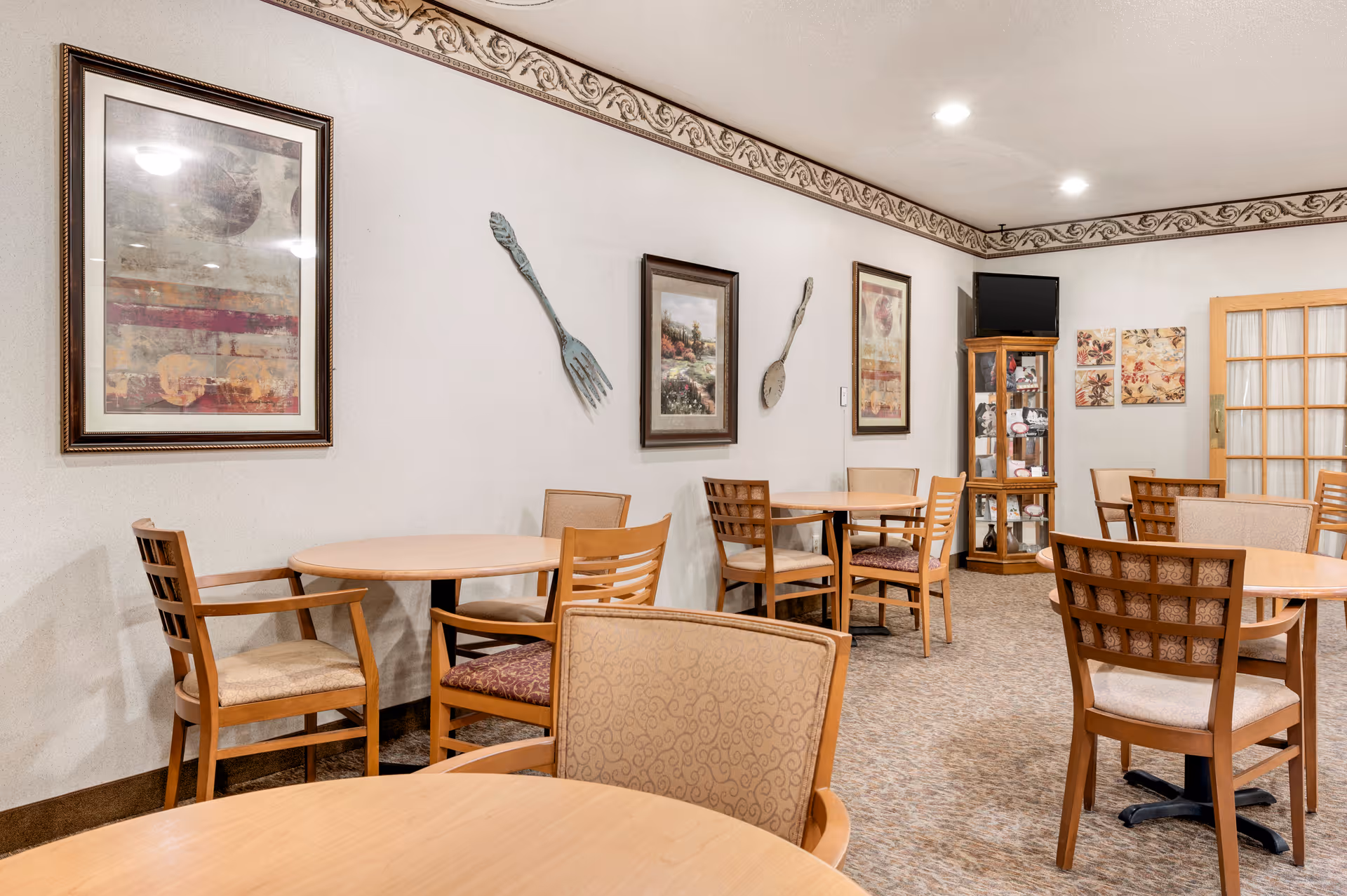 A dining room with several round wooden tables and chairs arranged around the room. The walls are decorated with framed artwork and large decorative fork and spoon wall hangings. There is a wooden display cabinet with a small television on top in the corner, and a door with glass panels on the right side.