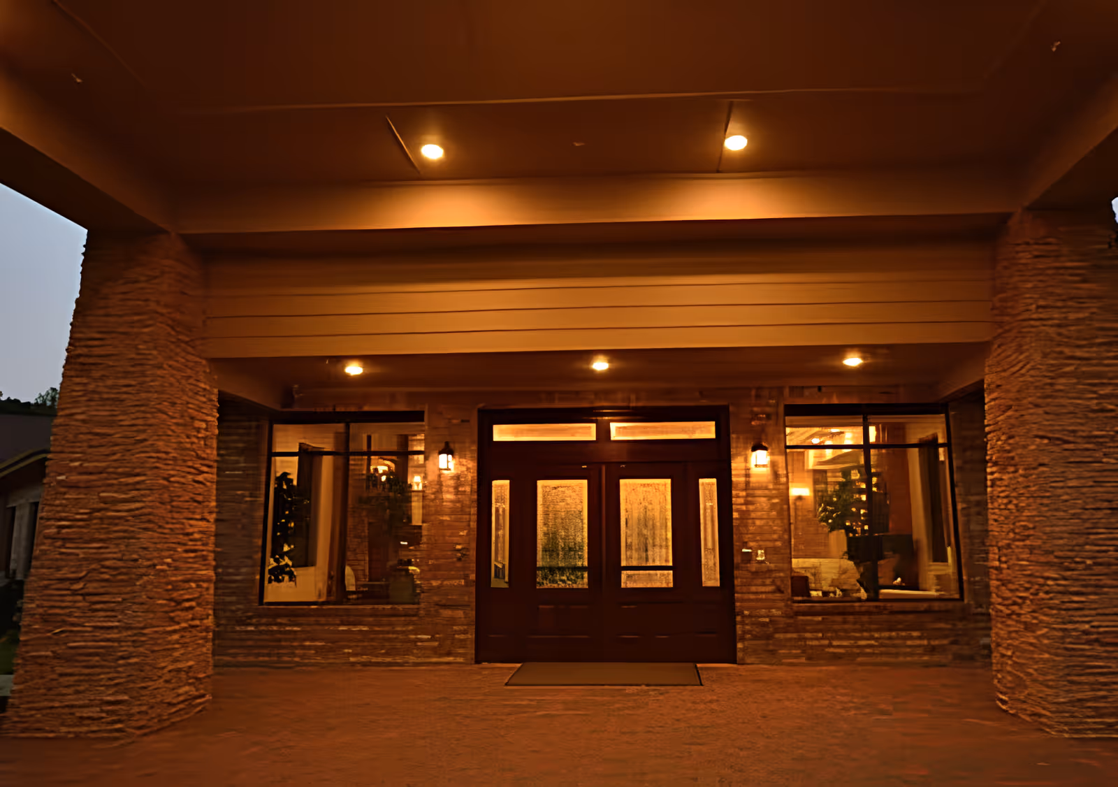 Entrance of a building at dusk with warm lighting, featuring a double wooden door with glass panels, flanked by large windows and stone pillars on either side.