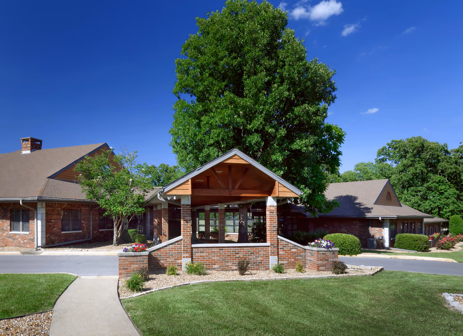 Exterior view of Butterfield Residential Care Center showing a brick building with a covered entrance, surrounded by green lawns, trees, and a clear blue sky.