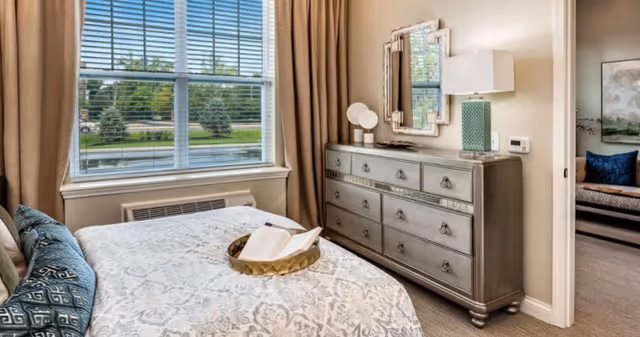 Bright bedroom featuring a patterned bed, a large dresser with mirror, and a window with blinds overlooking landscaped grounds.