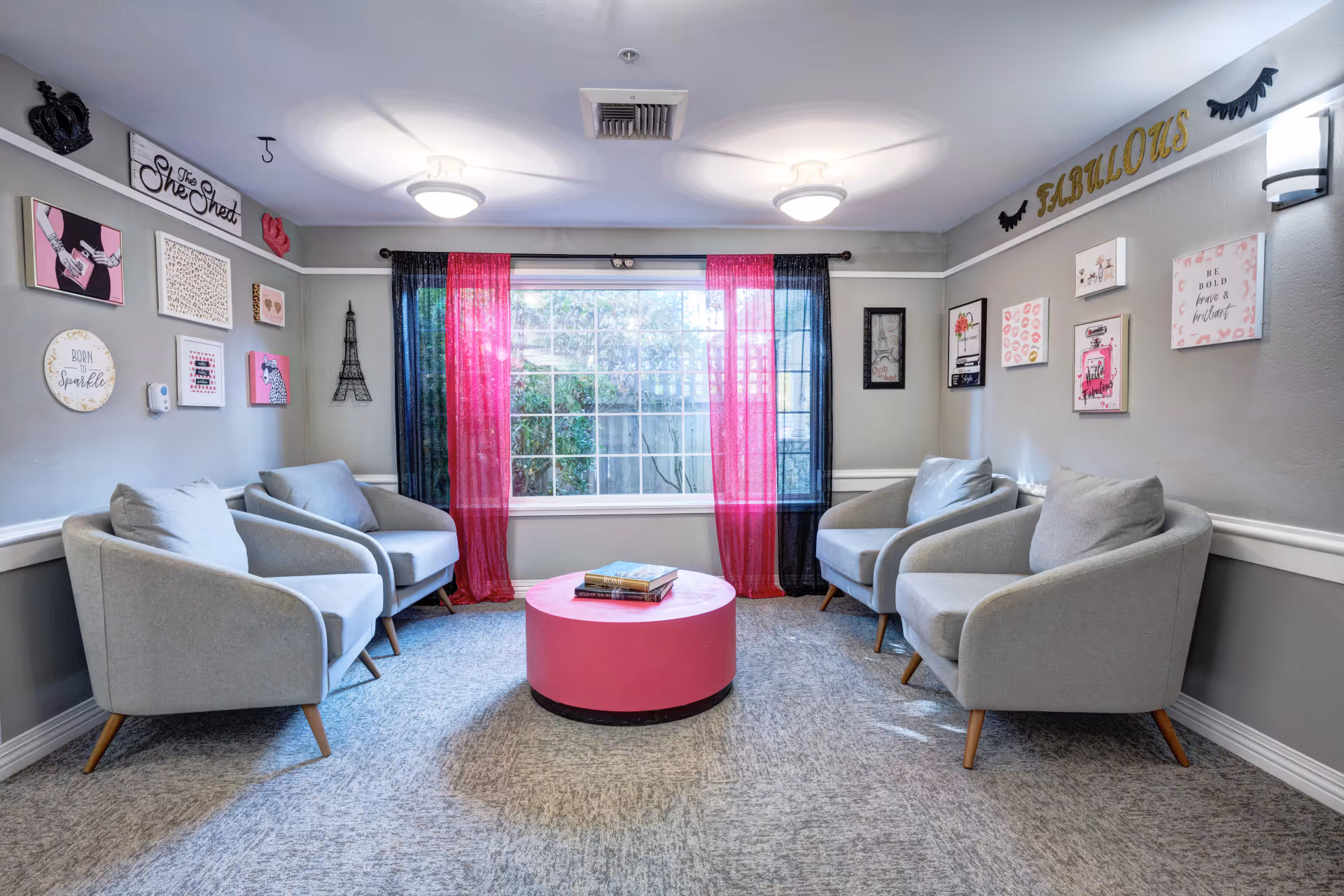 A cozy sitting area with four light gray armchairs arranged around a round pink coffee table with books on it. The room has gray walls decorated with various framed artworks and signs, including one that says 'The She Shed' and another that says 'FABULOUS'. A large window with sheer pink and black curtains lets in natural light, and the carpet is a light gray color.