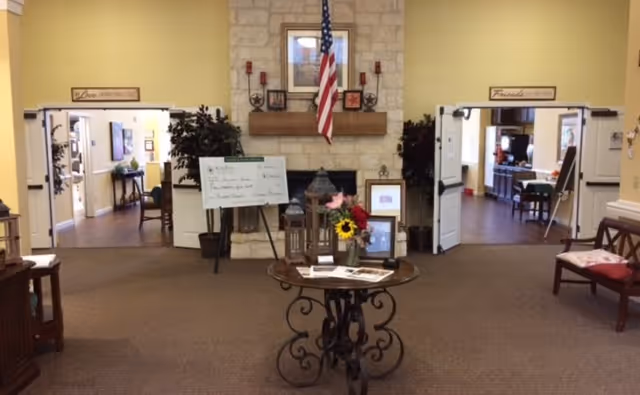Interior view of a senior living facility lobby area with a stone fireplace in the center, an American flag hanging above it, and a round table with decorative items including flowers and lanterns. Two open double doors on either side lead to other rooms with chairs and tables. The walls are painted yellow and there are framed pictures and plants around.