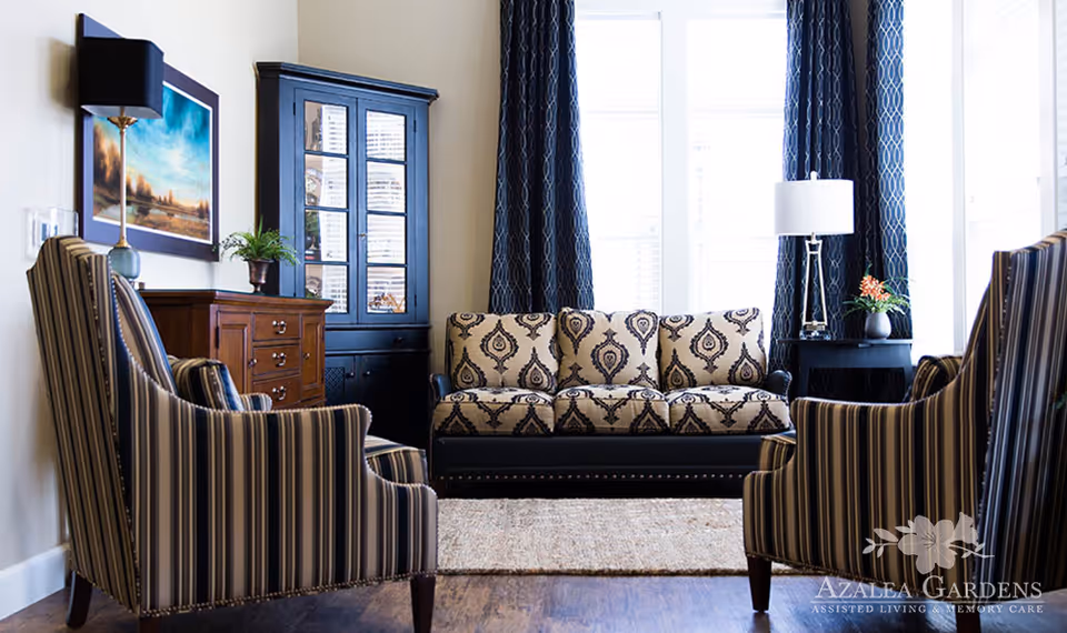 A bright and cozy living room with a patterned three-seater sofa, two striped armchairs, a wooden cabinet with glass doors, a side table with a lamp and plant, and large windows with dark curtains letting in natural light.