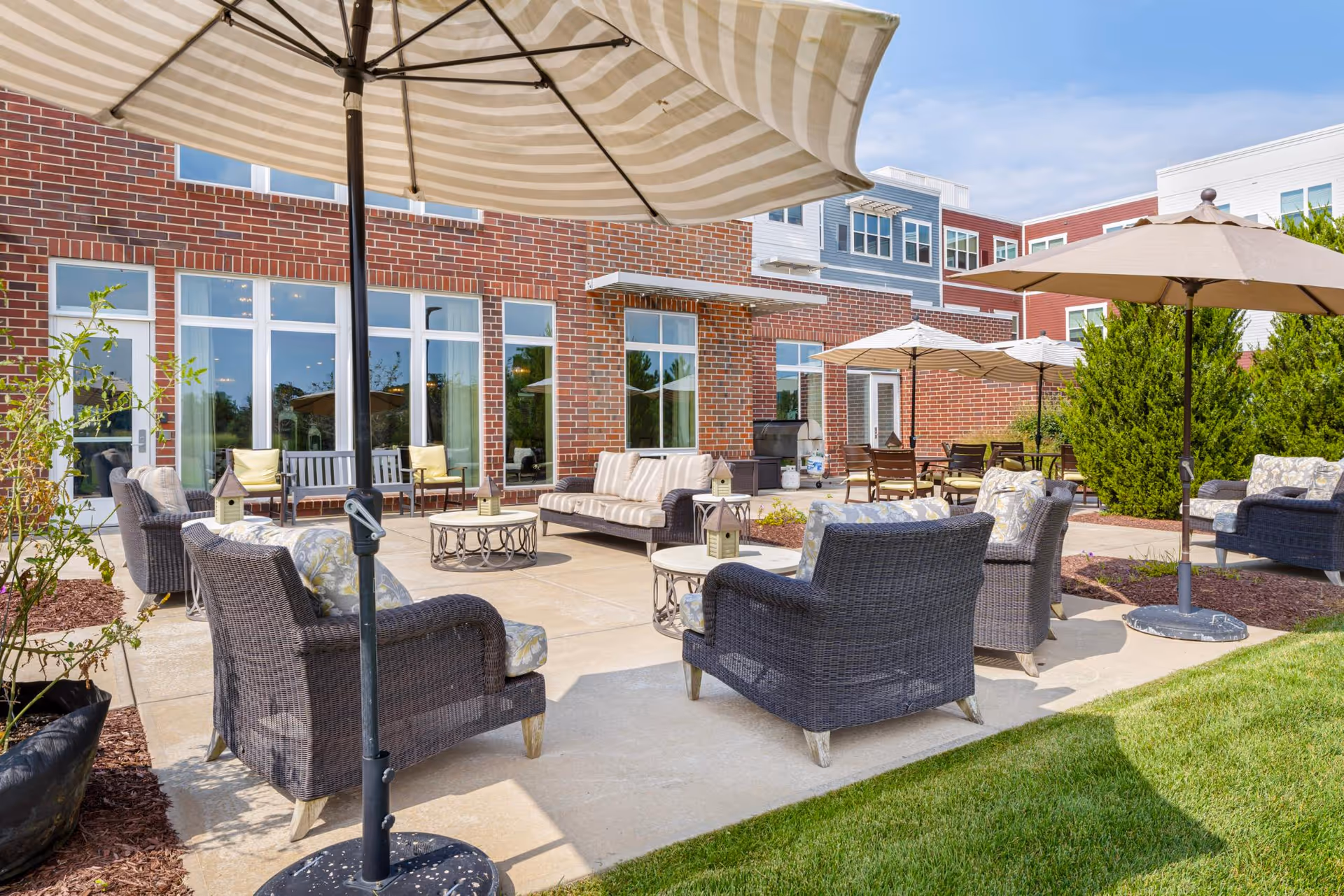 Sunlit outdoor patio with wicker chairs, sofas, umbrellas, and tables arranged on a concrete terrace in front of a brick building.