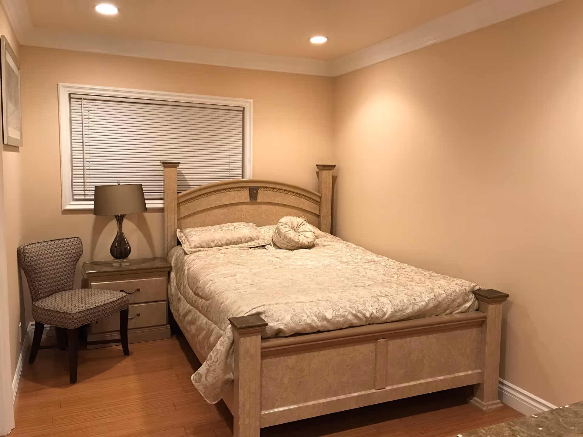 A bedroom with a beige color scheme featuring a large bed with a decorative headboard and matching bedding. Next to the bed is a nightstand with a dark lamp and a patterned chair. The room has wooden flooring, beige walls, a window with closed blinds, and recessed ceiling lights.
