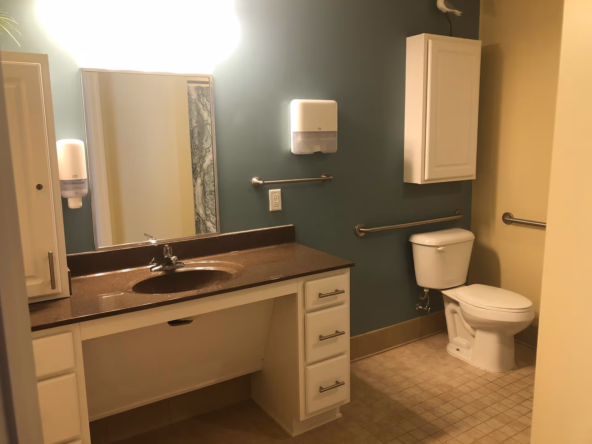 A clean and accessible bathroom with a white toilet, a brown countertop with an integrated sink, a large mirror above the sink, white cabinets, and grab bars on the walls for support. The walls are painted in two tones: teal and beige. There is a soap dispenser mounted on the wall next to the mirror.