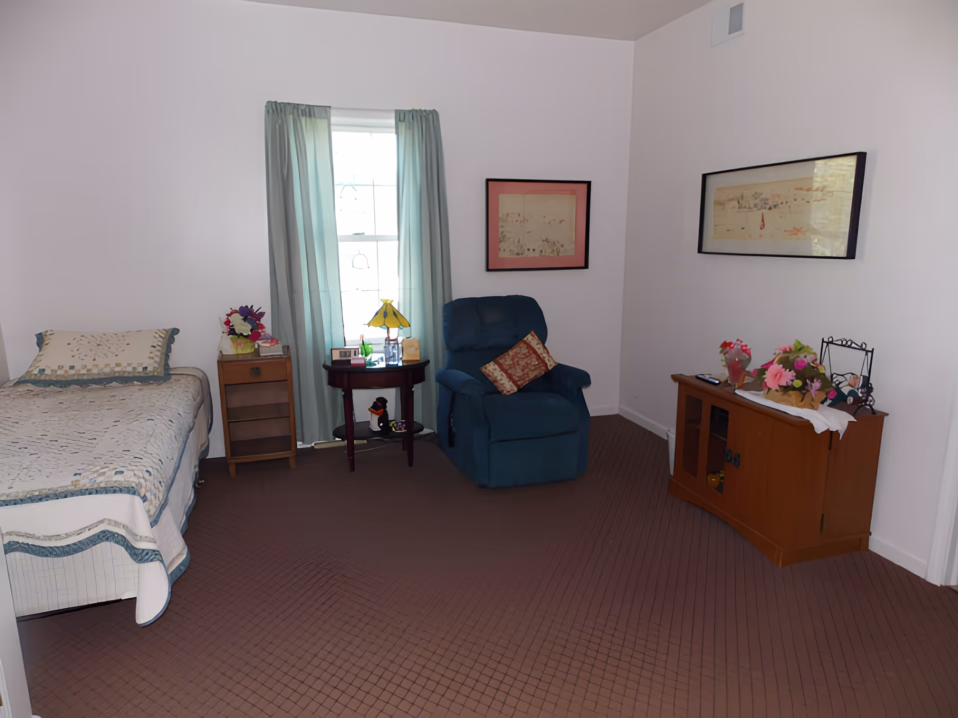 A cozy bedroom with a single bed covered with a quilt, a small wooden nightstand with a flower arrangement, a round table with a yellow lamp and decorative items, a blue recliner chair with a patterned cushion, and a wooden cabinet with flowers and framed pictures on top. The room has light-colored walls, a window with light blue curtains, and two framed artworks on the walls.