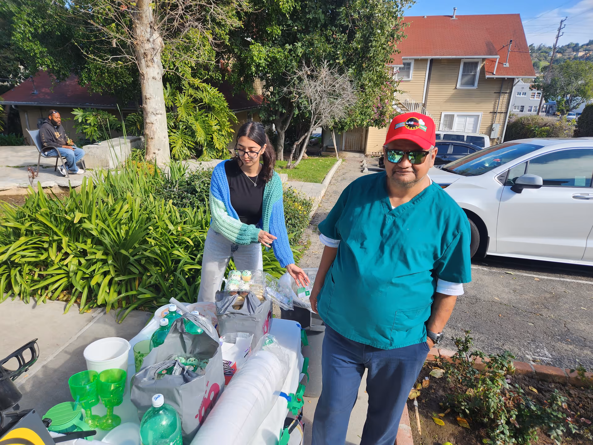 Two people outdoors near a table with green plastic cups, bottles, and food items. One person, wearing a red cap, sunglasses, and green scrubs, stands facing the camera. The other person, wearing glasses and a blue and green sweater, is arranging items on the table. In the background, a man sits on a bench near greenery and a tree, with houses and parked cars visible.