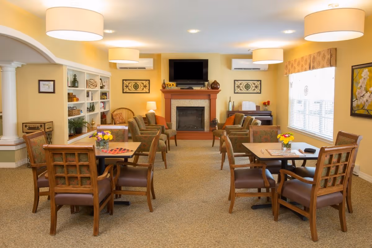 A cozy senior living common area with two square tables each surrounded by four wooden chairs with cushioned seats. The room features a fireplace with a TV mounted above it, several armchairs arranged around the fireplace, a piano in the corner, built-in shelves with decorative items, and large windows letting in natural light. The walls are painted a warm yellow, and the room is softly lit by ceiling lights and a table lamp.