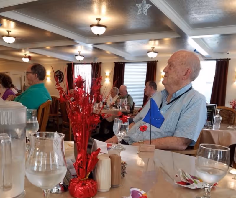 A group of elderly people sitting at tables in a dining room with red decorative centerpieces and glasses of water. The room has ceiling lights, windows with dark curtains, and a calm atmosphere.