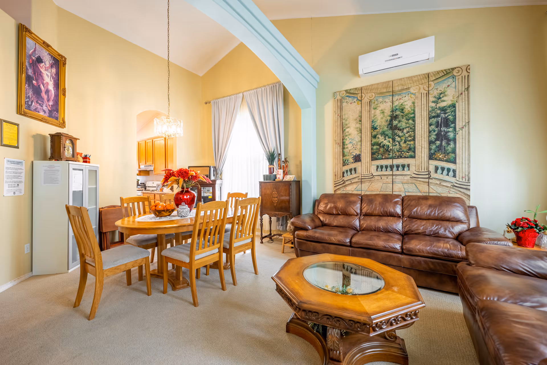 A bright and cozy senior living area featuring a brown leather sectional sofa, an ornate wooden coffee table with a glass top, and a wooden dining table with six chairs. The room has light yellow walls, a large window with white curtains, a decorative wall panel depicting a garden scene with columns, and a chandelier hanging above the dining table. There is also a white cabinet with papers posted on it and a framed painting on the wall.