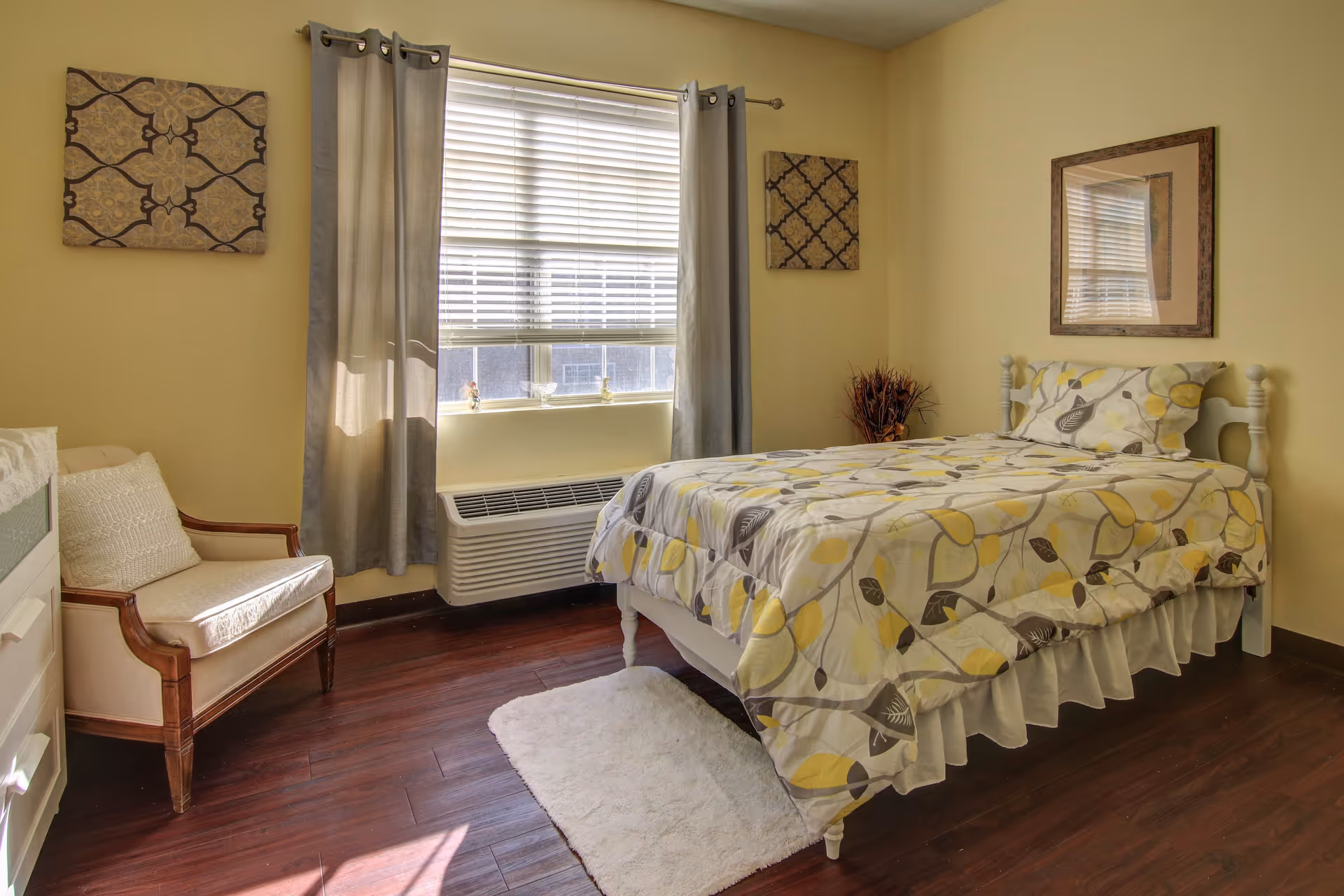 Sunlit bedroom with a single bed in yellow-gray patterned bedding, an armchair, window with blinds and curtains, and wall decor.