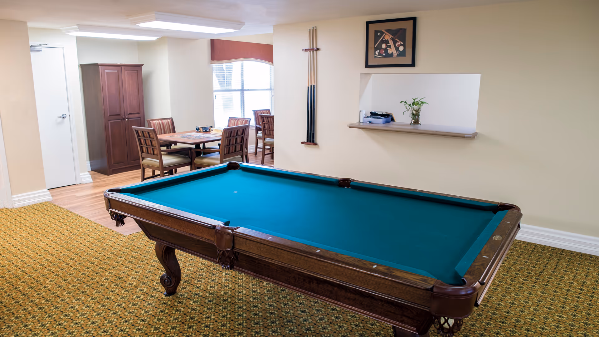 A recreational room with a green felt pool table in the foreground. Behind it, there is a small dining area with a table and four chairs near a window. Pool cues are mounted on the wall next to a framed picture of billiard balls and cues. A small shelf with a plant and some items is built into the wall.
